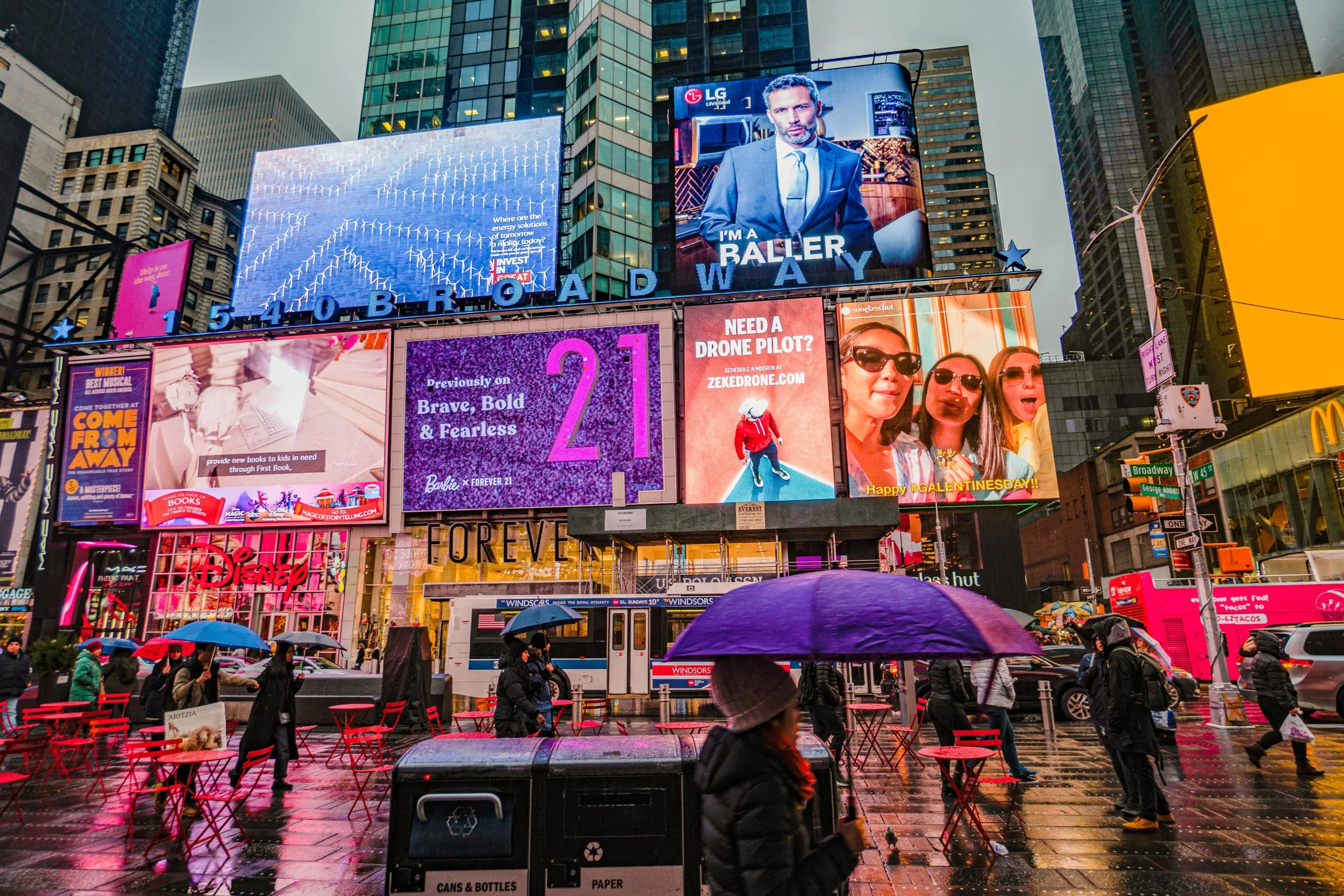 Colorful digital billboards in a busy city street with people walking under umbrellas on a rainy day.
