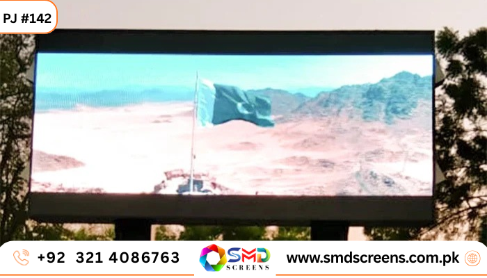 A large outdoor LED billboard or screen is shown displaying a scenic image of a Pakistani flag (green with a white stripe and a crescent moon and star) flying on a flagpole in a vast desert or mountainous landscape.