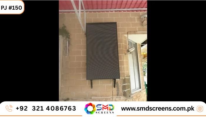 A large, tall outdoor LED screen (currently off/dark) mounted vertically on a light brown brick wall next to a window.